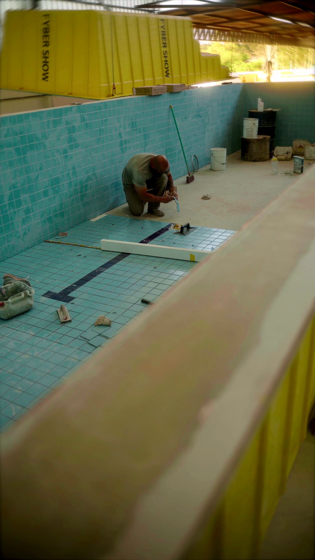 Funcionário ajoelhado no fundo da piscina conferindo o alinhamento das pastilhas, com moldes amarelos da Fyber Show ao fundo
