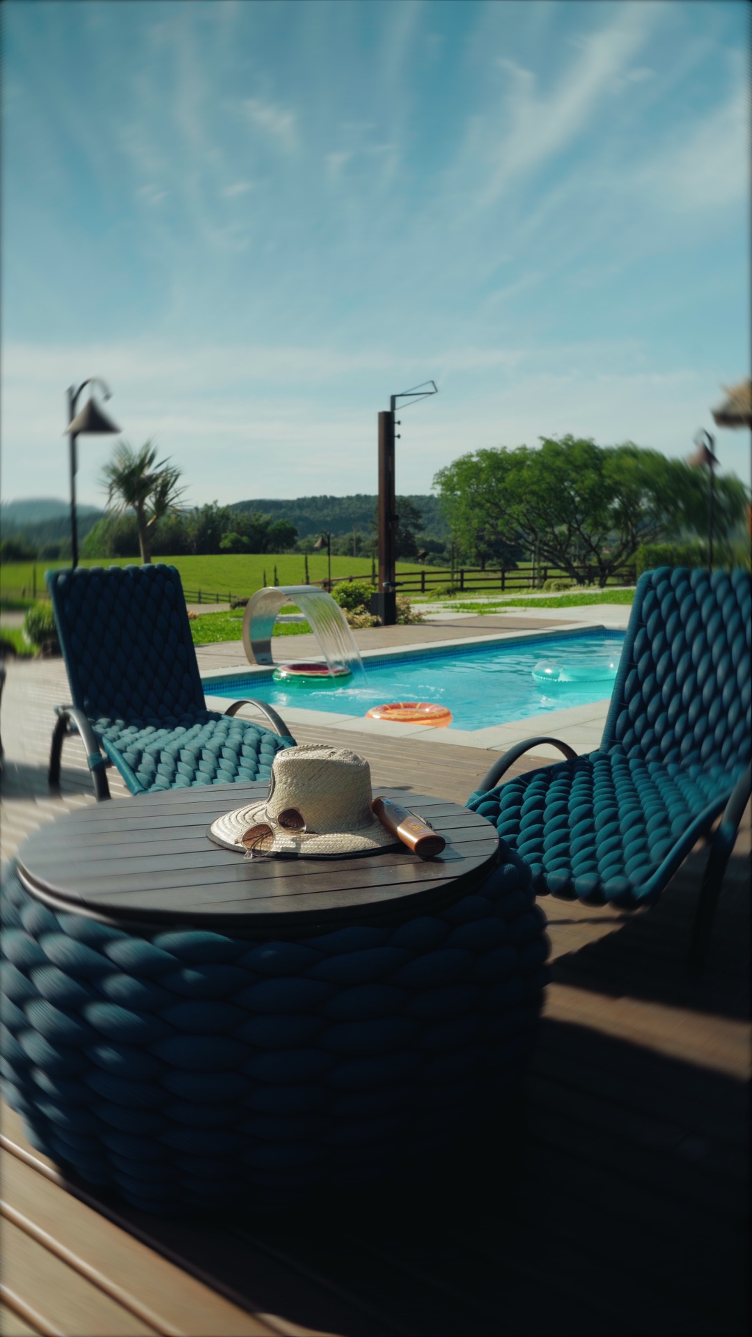 Deck de madeira com duas espreguiçadeiras e uma mesa, a piscina com cascata ao fundo e o campo atrás