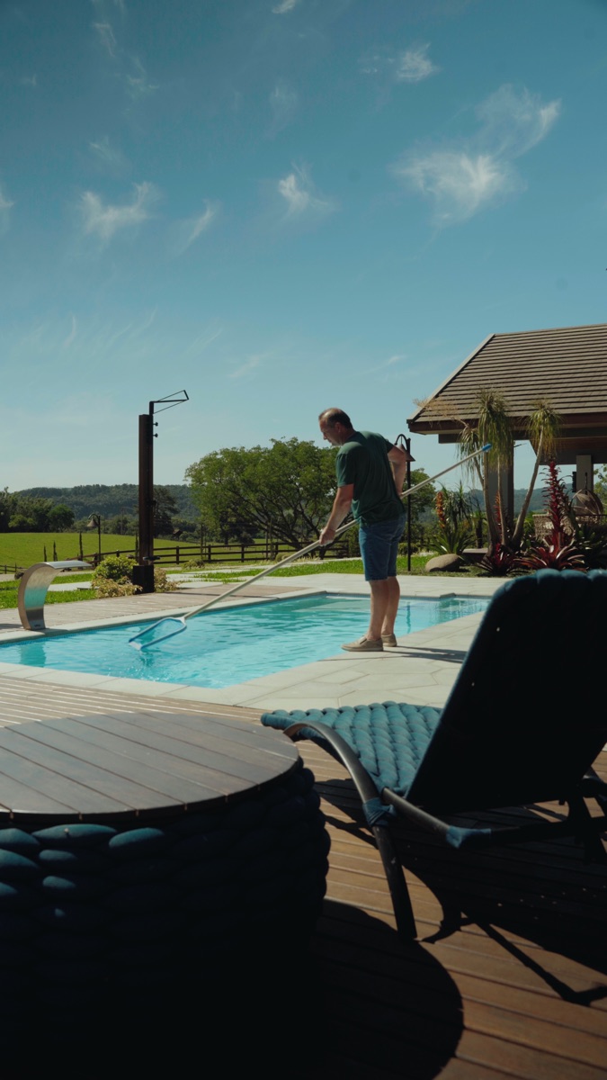 Homem limpando a piscina com o cabo de manutenção, no deck, ao entardecer